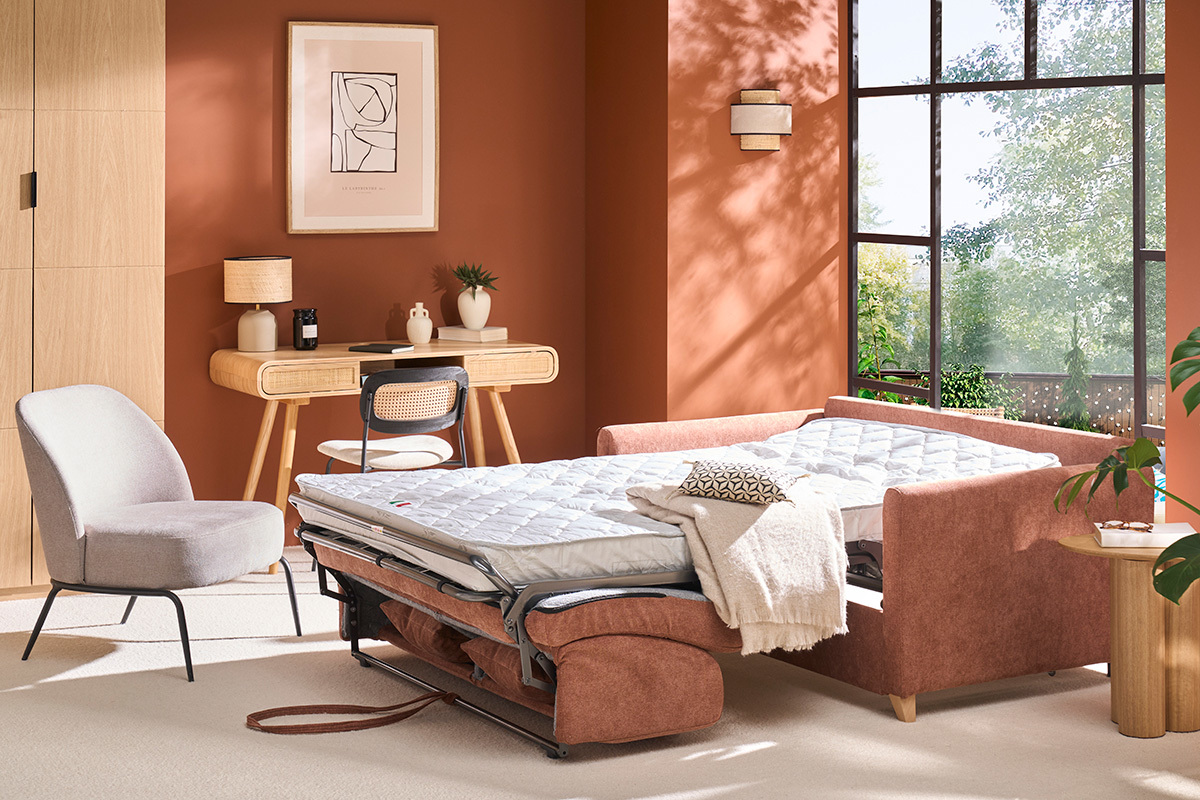 Canap� convertible en tissu terre br�l�e ouvert en lit avec matelas, table et chaise en fond.