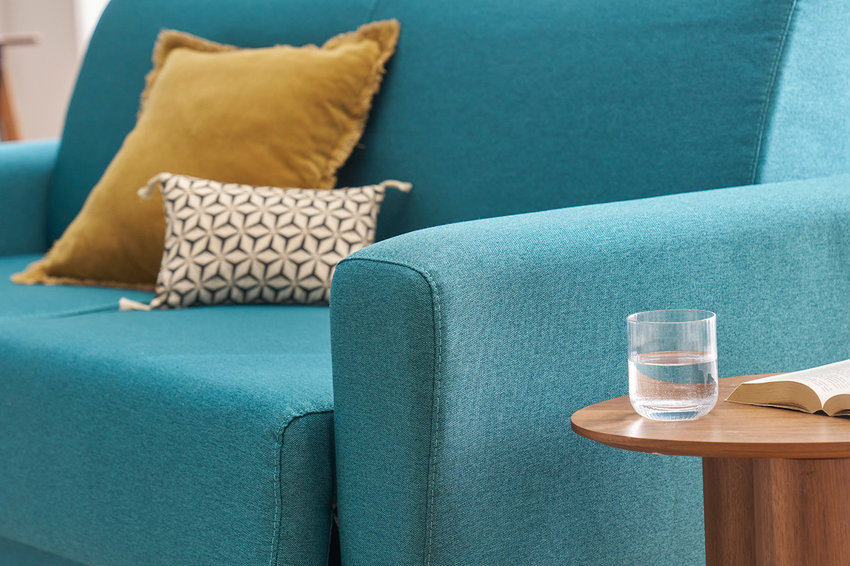 Canap� convertible bleu canard avec coussins sur une table d'appoint en bois et un verre d'eau.