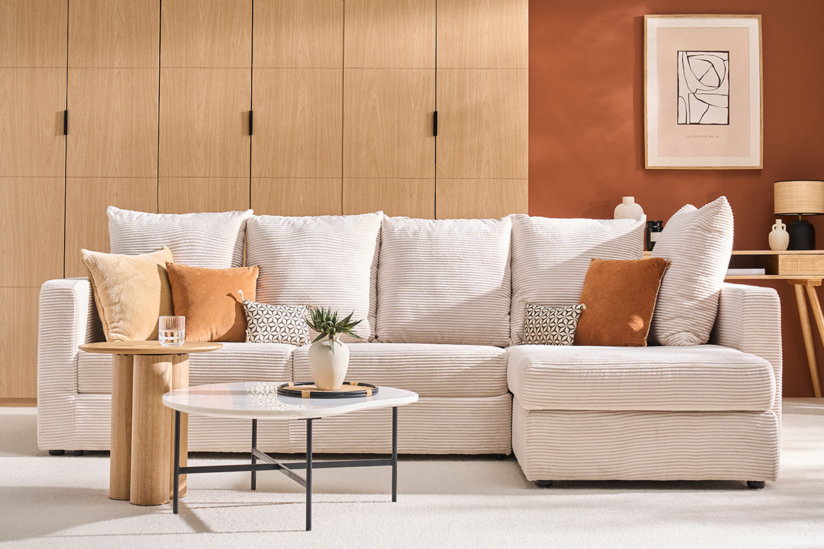 Canap� d'angle convertible en velours c�tel� beige, coussins assortis, table basse et plante, fond bois clair.