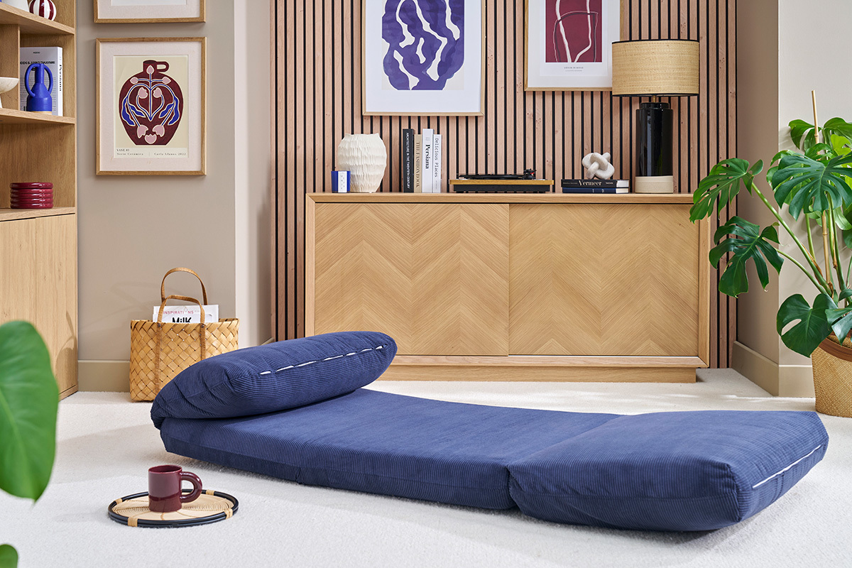 Chauffeuse en velours c�tel� bleu fonc� SPENCER d�pli�e sur tapis, avec meuble en bois et plantes en arri�re-plan.