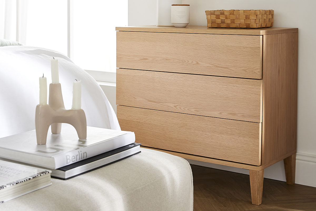 Commode scandinave en bois clair � 3 tiroirs pr�s d'un lit, vue de c�t�.