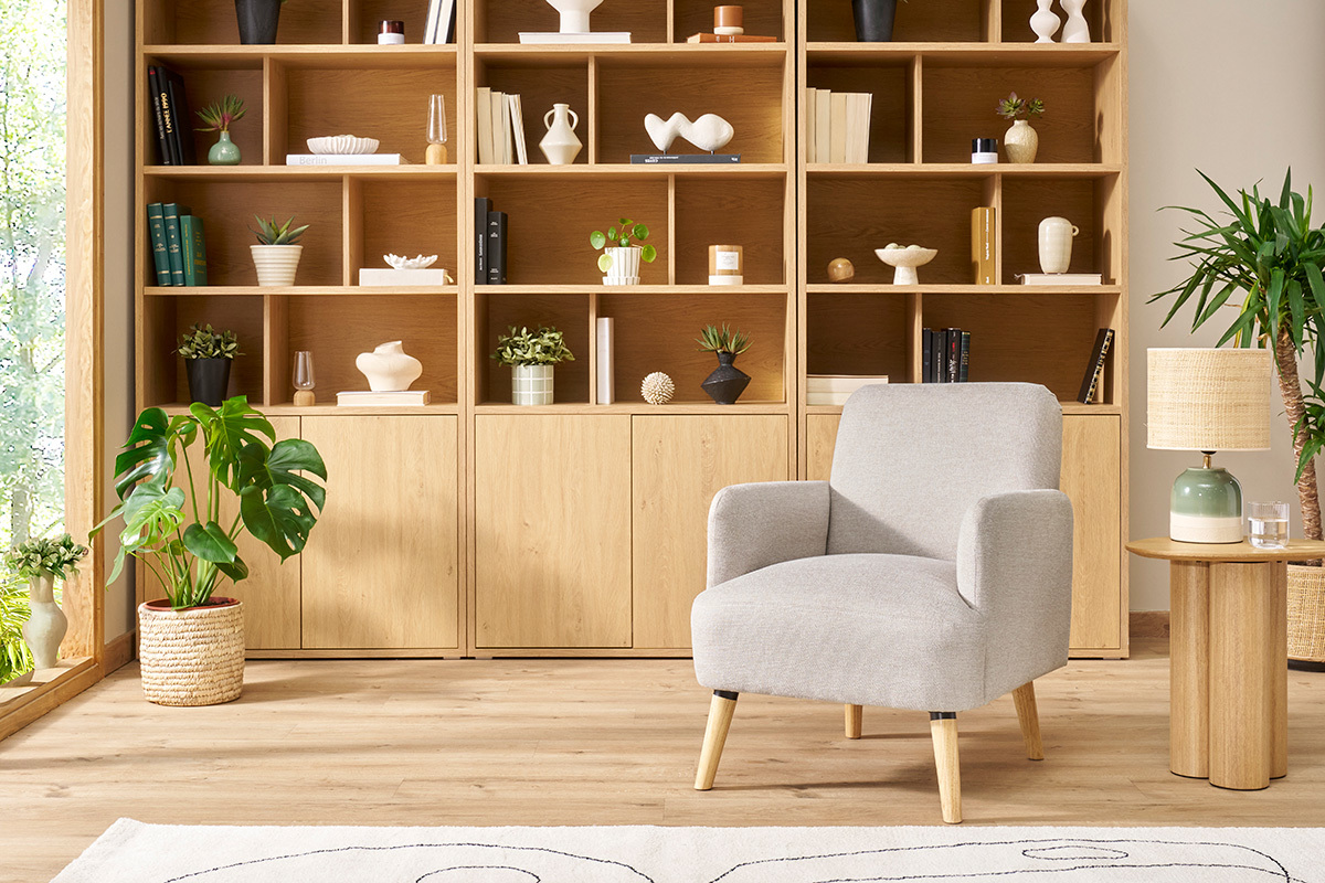 Fauteuil ISKO beige et en bois clair pos� dans un salon avec �tag�res et plantes.