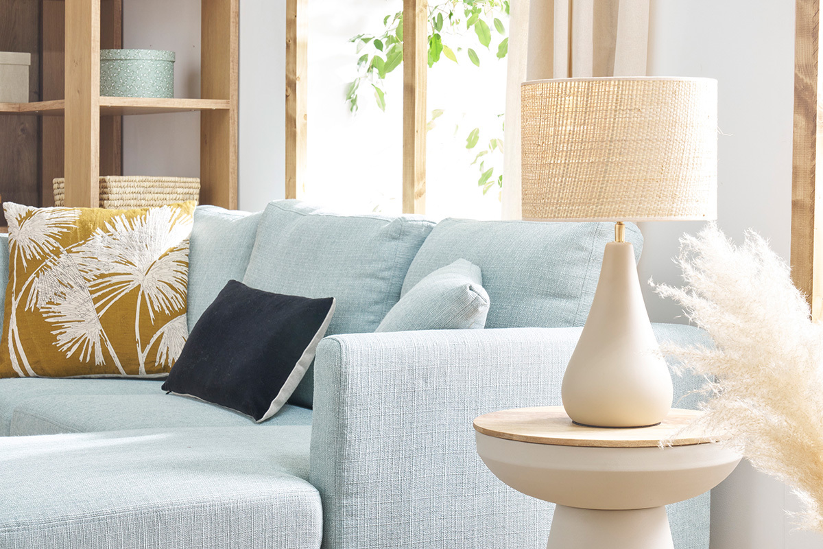 Lampe � poser taupe en c�ramique avec abat-jour en raphia sur table � c�t� d'un canap� bleu.
