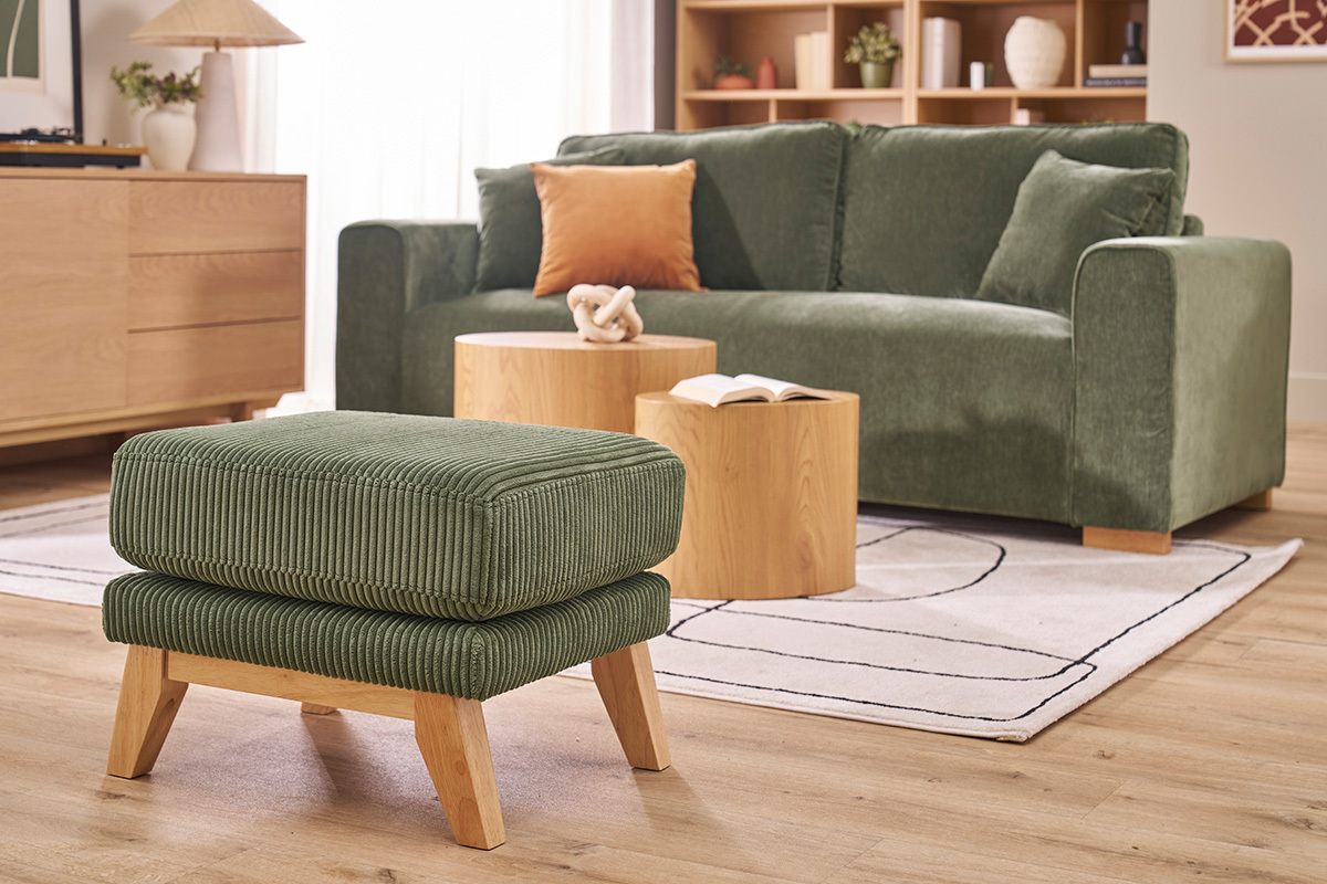 Pouf en velours c�tel� vert kaki avec pieds en bois clair devant un canap�, vue de profil.